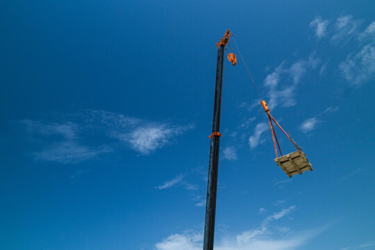 Large construction mobile crane lifting a heavy wooden cargo pallet high up into the bright blue sky with wispy white clouds during a commercial building project operation at a job site