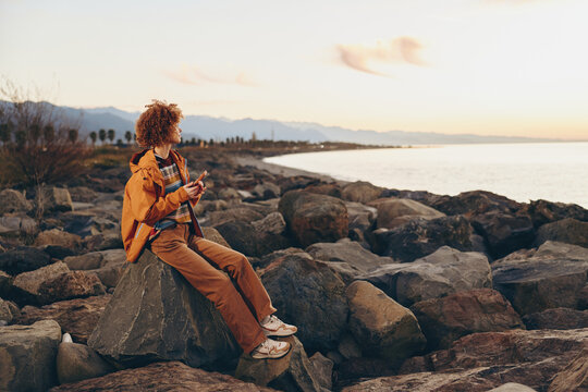 Woman lifestyle on seaside rocks at sunset, coastal ocean waves and warm sky. She sits on the shoreline in an orange jacket, travel mood, outdoor scene with horizon and calm water.