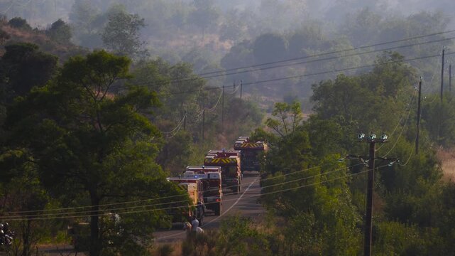 Dramatic telephoto shot of a convoy of French firefighting trucks (CCF), flashing blue lights, driving down a narrow road enveloped in thick smoke, heading toward a forest fire near Salagou Lake.