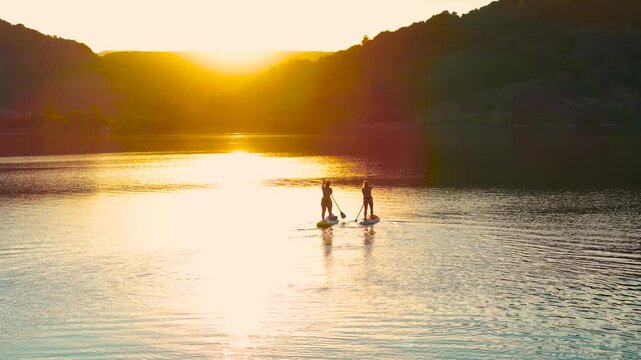 Aerial drone footage capturing a closer view of two people on stand-up paddleboards (SUP) paddling together on the calm, golden-lit waters of Salagou Lake (Lac du Salagou) during a warm summer sunset.