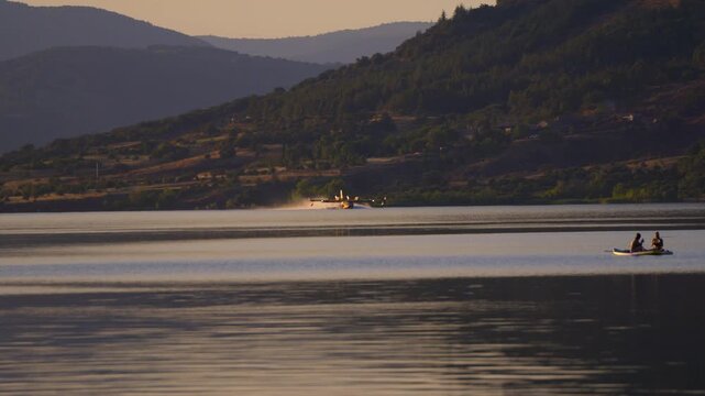 Dramatic telephoto aerial shot of a firefighting Canadair aircraft scooping water and leaving a massive mist spray on Salagou Lake (H&eacute;rault, France), with two people on a paddleboard observing action