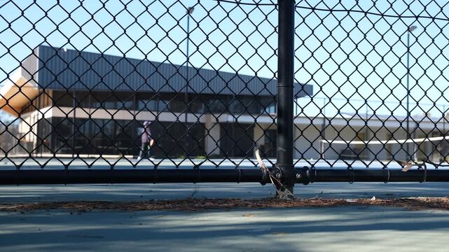 A black chain-link fence onto an outdoor suburban tennis court in Melbourne, Australia, with a blurry view of teenagers play recreational tennis in front of a modern community sports pavilion.&nbsp;