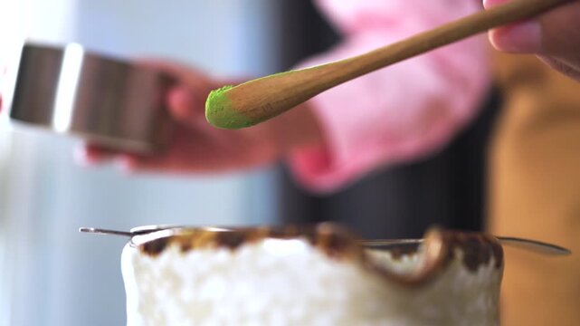 Close-up of hands using a wooden scoop to sift organic matcha green tea powder through a metal strainer into a ceramic bowl. Japanese tea ceremony and cafe culture concept.