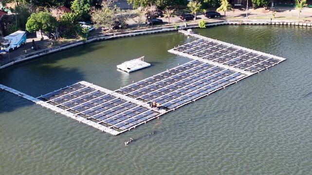 Aerial view of the PLN Indonesia Power floating solar panels on the Muara Nusa Dua reservoir, with surrounding vegetation and calm water in Kecamatan Denpasar Selatan, Bali, Indonesia.