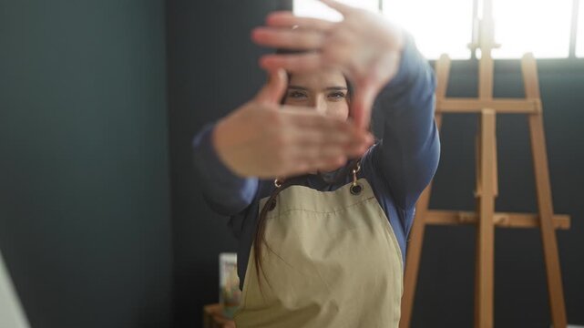 Woman frames view with hands forming a rectangle, wearing an apron beside an easel in studio; contemplative focus.