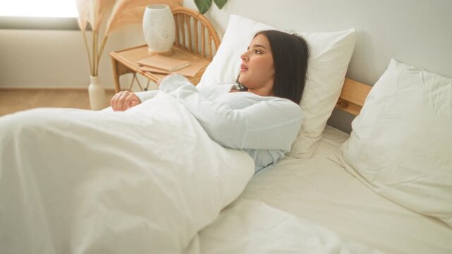Woman sits up on bed pulling long hair back while tucking white duvet beside rattan headboard and potted plant near window in bright studio; serenity.