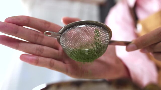 Close-up of hands tapping a metal strainer, sifting fine organic matcha green tea powder falling into a ceramic bowl. Japanese tea making process and healthy cafe beverage concept.