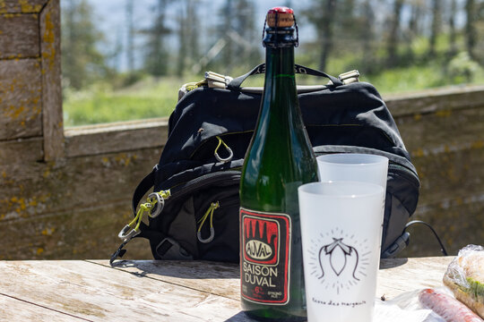 Gaspesie, Quebec, 15 July 2026 : A bottle of Auval Saison Duval strong ale, two plastic cups, and a backpack rest on a wooden table outdoors.