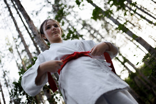 Young girl tying a red belt in a karate gi outdoors in a forest during martial arts training and focused practice