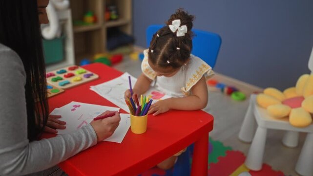 Woman teacher assists little girl drawing at a red table, hand holding crayon and paper visible in a building; concentration early education.