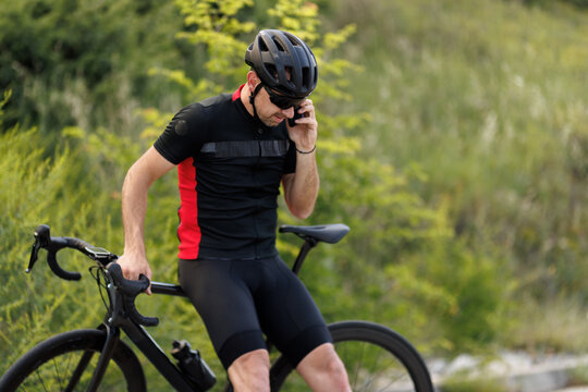 Cyclist resting on road bike while talking on phone in outdoor location with greenery in the background