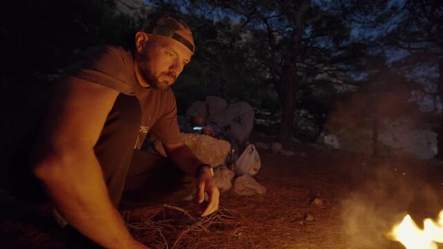 man sitting and interacting with a campfire in a relaxed and contemplative outdoor nighttime setting near a campfire with wooden firewood, close-up slightly low angle. High quality 4k footage