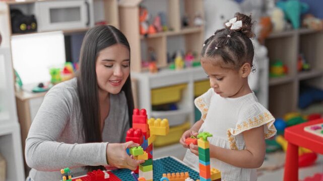 Woman guiding little girl as they stack colorful interlocking toy blocks on a play table in daycare building; nurturing play.