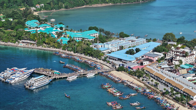Aerial view of Koh Phi Phi showing the pier with numerous long-tail boats, turquoise water, and coastal resort buildings in Khlong Phon, Krabi, Thailand.