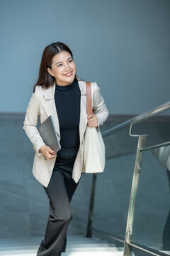 Smiling businesswoman walking up stairs in an office building with a laptop and shoulder bag, dressed in smart work clothes