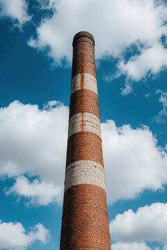 Old red brick industrial chimney stands against bright blue sky with white clouds. Tall vertical factory pipe rises from ground level showing aged masonry texture. Historic power station tower