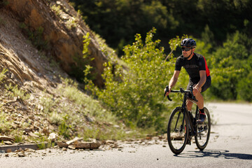 Fototapeta premium Cyclist rides a road bike on a sunny day along a winding mountain road surrounded by greenery and rocky terrain