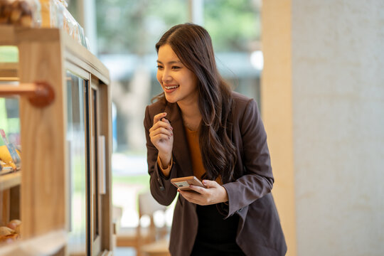 Smiling young asian woman using a smartphone while browsing a cafe display case indoors, with warm light and a relaxed shopping moment.