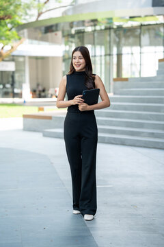 Young businesswoman standing outdoors with a tablet near a modern office building, smiling confidently in a bright urban setting.