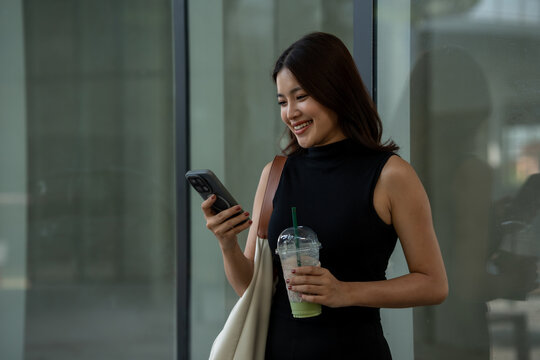 Smiling young woman in a black dress checking her phone while holding an iced drink and tote bag outside a modern office building.