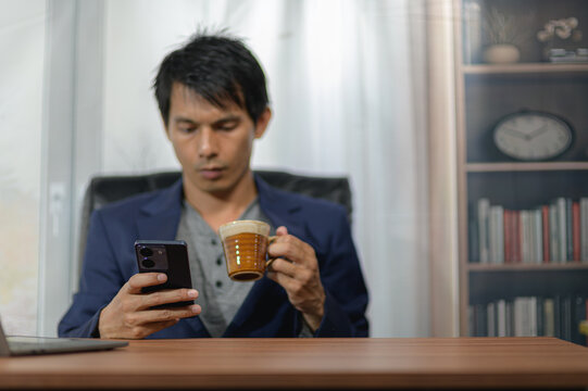 Young man with black hair in blue blazer using smartphone and drinking coffee at desk in home office