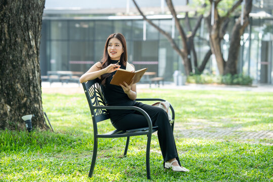 Young woman sitting in a green park, holding a notebook and smiling in sunlight, relaxing and reading outdoors