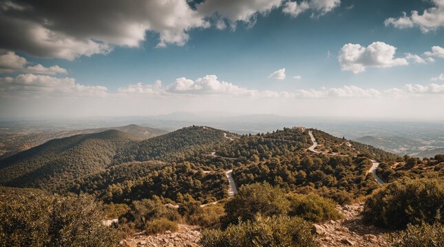 Scenic view from Mount in Cyprus. Winding road snakes through dense pine forest covered hills under bright blue sky with white clouds. Distant horizon fades into haze. Natural outdoor landscape