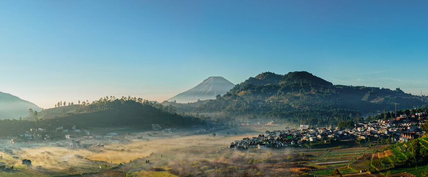 Aerial view of the misty Dieng Plateau landscape featuring a small village nestled among volcanic mountains and terraced fields under a clear blue sky in Indonesia.