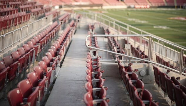 Medium shot of a lower bowl grandstand section showcasing rows of deep red seats with aisles and railings the field partially visible in soft focus beyond.