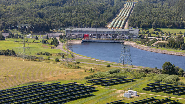 Aerial view of a hydroelectric power plant with large water pipes, a reservoir, solar panel arrays, and high-voltage electricity pylons set in a rural landscape near Gdansk, Poland.