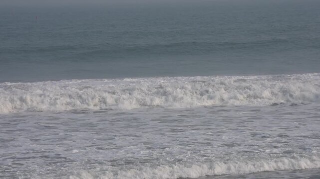 Waves gently roll onto the shore near Yogyakarta, Indonesia, creating white foam against dark blue ocean waters in a calm coastal scene.