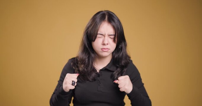 Young woman feeling strong emotions, clenching fists and grimacing against plain background