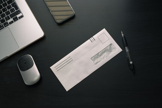 A top-down view of a white envelope, a pen, a wireless mouse, a phone, and a laptop arranged on a black office desk.
