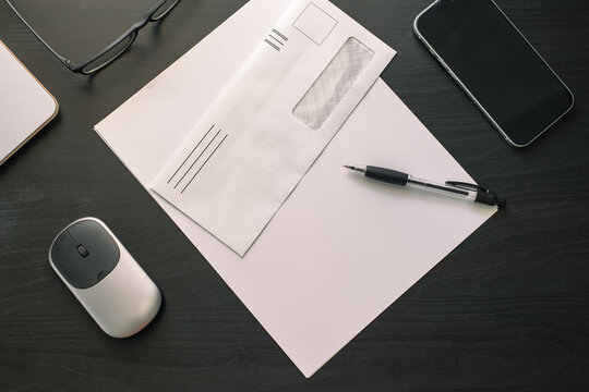 A flat lay of a white window envelope, a black pen, a wireless mouse, a phone, and eyeglasses on a dark wooden desk surface.