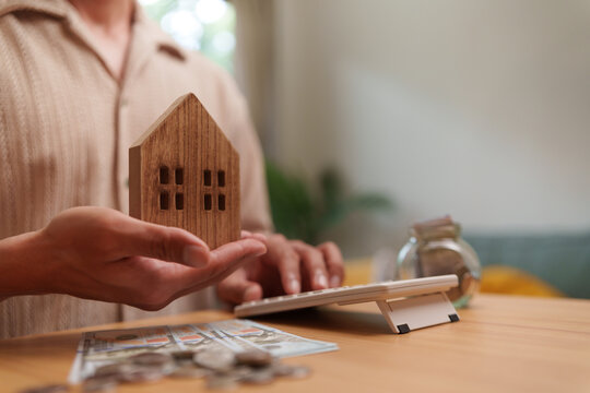 Man calculating home investment costs while holding a wooden house model, planning mortgage, savings, and future finances.