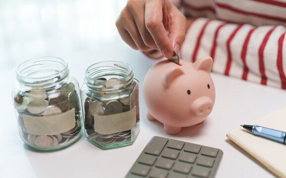 Hand placing a coin into a piggy bank beside savings jars, representing personal finance, emergency savings, retirement planning, and disciplined money management.