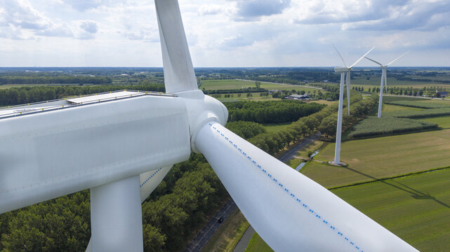 Aerial view of large white wind turbines set amidst lush green fields, trees, and a road in the rural landscape of Culemborg, Gelderland, Netherlands.