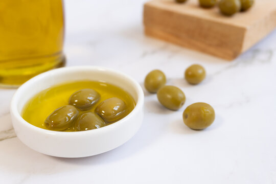 Fresh green olives soak in golden olive oil. White bowl holds rich, aromatic oil. Marble surface adds clean, elegant contrast. Olives scattered nearby enhance natural appeal. Ideal for food, health