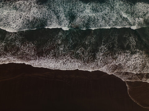 Aerial view of ocean waves crashing onto a dark sandy beach in Yogyakarta, Indonesia, with white foam and deep blue water.