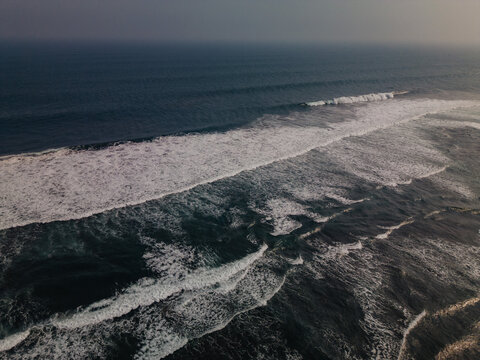 Aerial view of ocean waves crashing on a rocky shore, with foamy white water contrasting against dark blue sea.