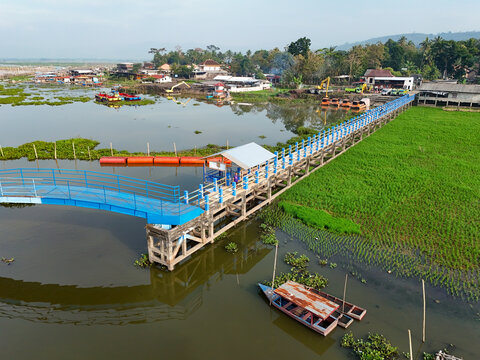 Aerial view of Jembatan Biru Rawapening stretching across the lake with lush green rice fields, small wooden boats, and water hyacinths in Kecamatan Bawen, Jawa Tengah, Indonesia.