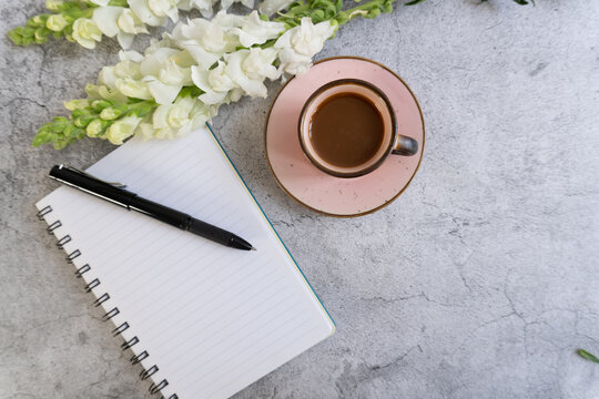 White flowers rest beside a coffee cup and open notebook. Black pen lies on lined pages, ready for writing. Pink saucer holds warm, dark beverage.