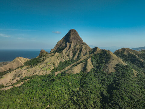 Aerial view of Nuca Molas mountain with rugged slopes and lush green vegetation against a clear blue sky and the ocean in Satar Mese Barat, East Nusa Tenggara, Indonesia.