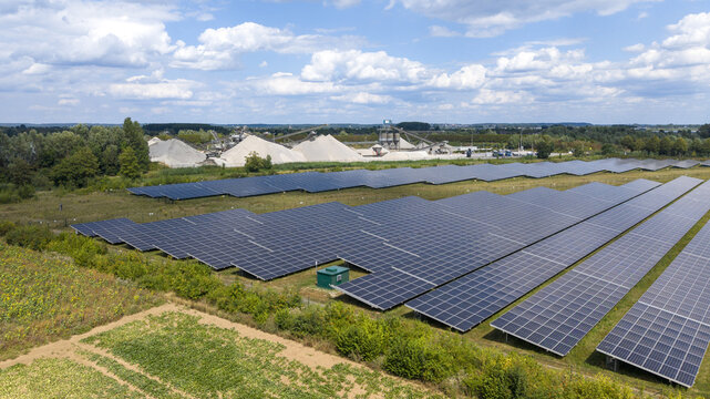 Aerial view of a solar farm with rows of photovoltaic panels in a rural landscape, with industrial gravel piles in the background under a blue sky in Echteld, Gelderland, Netherlands.