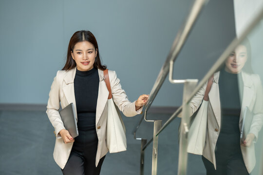 Businesswoman walking up indoor stairs with laptop and bag, seen with reflection in glass railing.