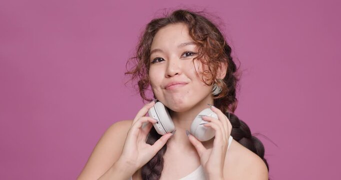 Young Kazakh woman enjoying music with white headphones against a pink background
