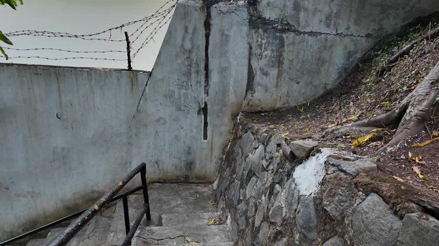 Narrow concrete pathway with stairs and stone retaining wall enclosed by security fencing.