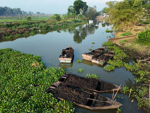 Aerial view of wooden boats filled with dark sediment on Rawa Pening waters surrounded by green water hyacinths and rural landscape in Waduk Rawapening, Central Java, Indonesia.