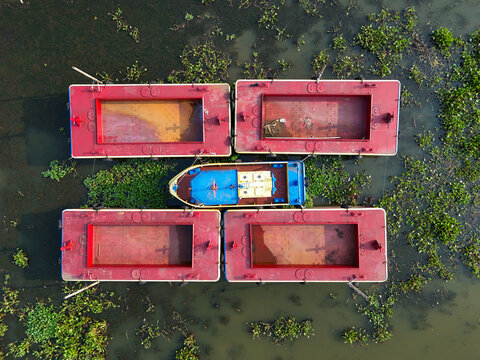 Aerial view of a blue boat docked among four large red rectangular floating platforms surrounded by water hyacinths in Waduk Rawapening, Central Java, Indonesia.