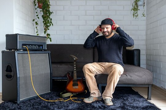 Hispanic musician sitting on couch to place on red headphones, electric guitar and amp stack background